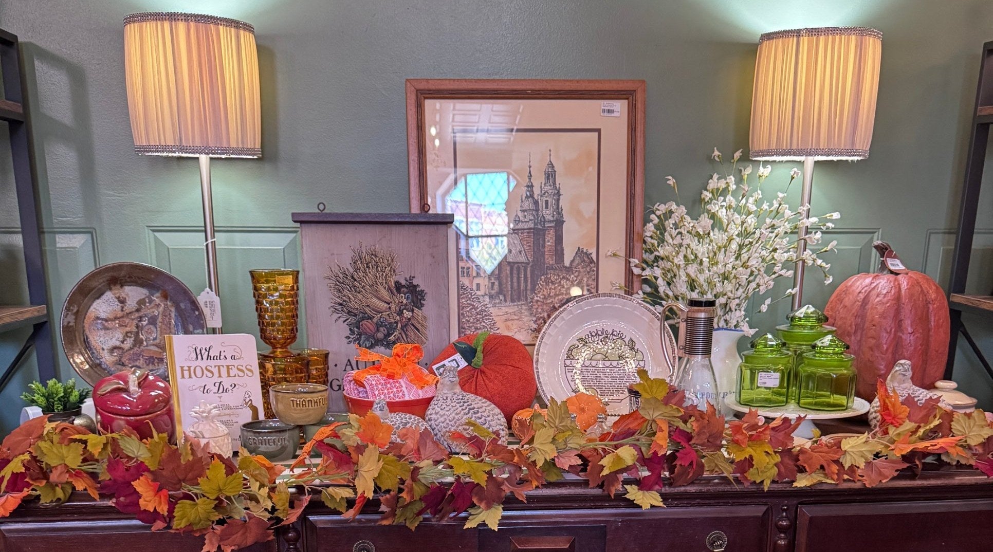 Thanksgiving display on a vintage wood breakfront at Jackson Square Collective, with fall leaf garland, pumpkins, amber glassware, green glass canisters, a harvest wheat sign and framed art. | Jackson Square Collective in La Grange Park, IL