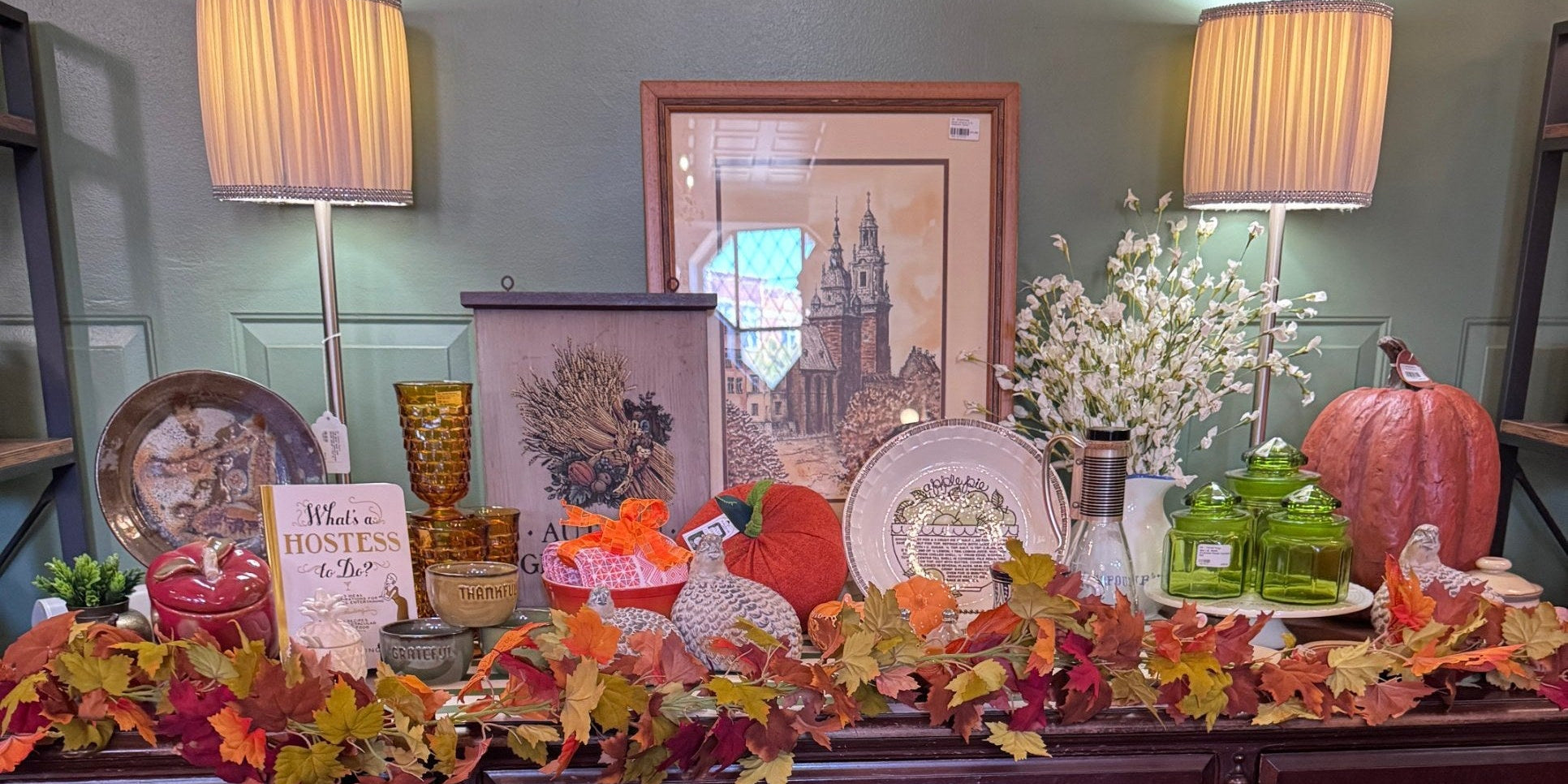 Thanksgiving display on a vintage wood breakfront at Jackson Square Collective, with fall leaf garland, pumpkins, amber glassware, green glass canisters, a harvest wheat sign and framed art. | Jackson Square Collective in La Grange Park, IL