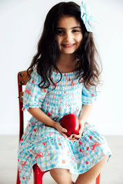Young girl in a patterned dress holding a red heart-shaped object, sitting on a chair against a white background.