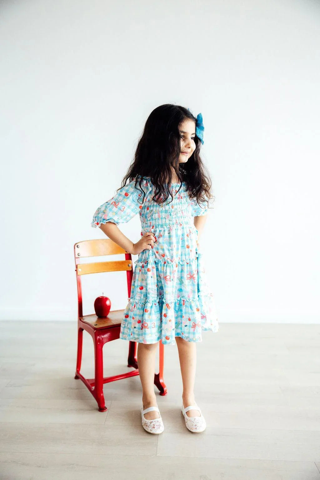 Young girl in a floral dress standing next to a chair with an apple on a white background