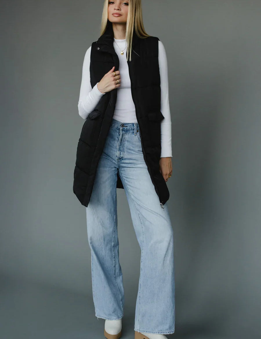 Woman wearing a black vest, white shirt, light blue jeans, and white shoes on a gray background – JSQ Mercantile Women’s Boutique & Gift Shop Located in La Grange, IL